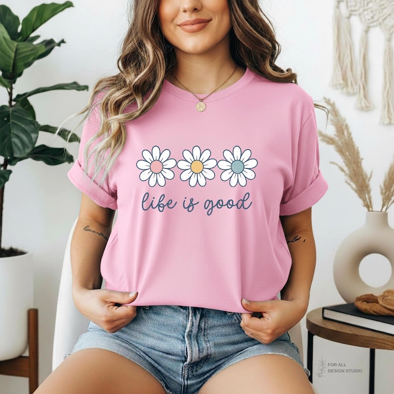 May include: A light pink t-shirt featuring three daisy flowers with pink, yellow, and blue centers. The words "life is good" are written in a cursive font below the flowers. The shirt is a casual style.