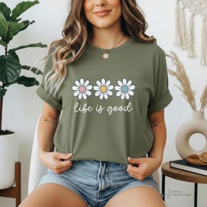May include: Olive green t-shirt featuring three white daisies with pastel centers and the phrase "life is good" in a cursive font. The shirt has rolled-up sleeves and is paired with denim shorts.