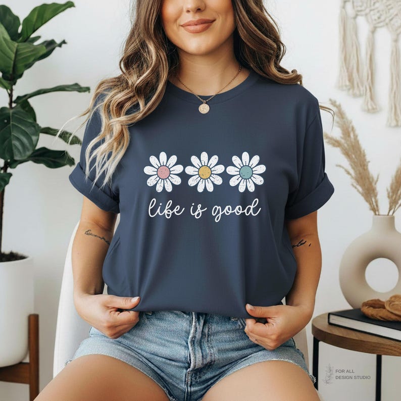 May include: A navy blue t-shirt featuring three daisy flowers with pastel centers and the phrase "life is good" in white script. The shirt has rolled-up sleeves and is worn with denim shorts.