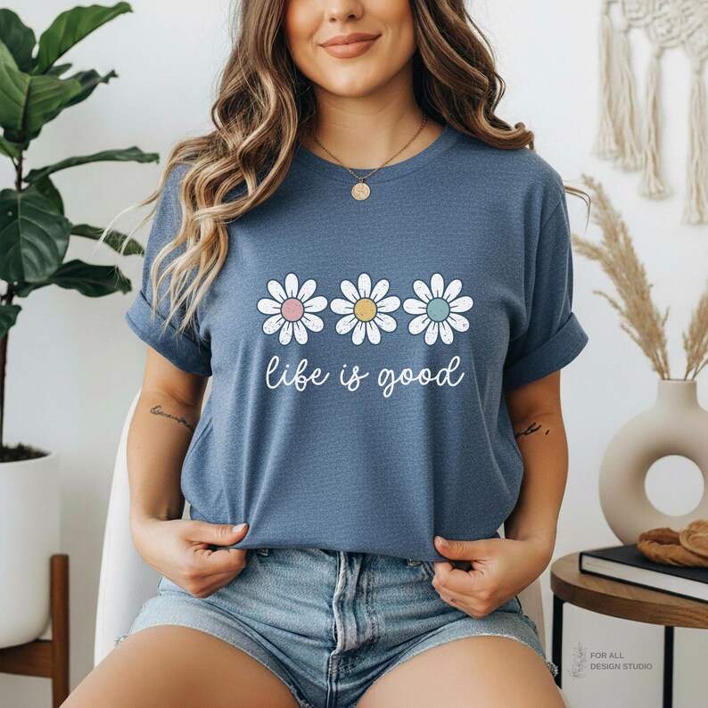 May include: A blue t-shirt with three daisy graphics in pastel colors and the phrase "life is good" in white script. The shirt is a casual style.