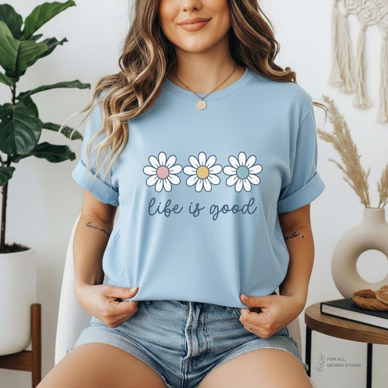 May include: Light blue t-shirt with three daisy graphics and the phrase "life is good" in a cursive font. The daisies have white petals with pink, yellow, and blue centers. The shirt has rolled-up sleeves.