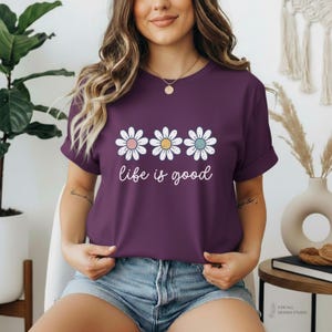 May include: A plum-colored t-shirt featuring three daisy graphics with pink, yellow, and blue centers. The words "life is good" are written in a cursive font below the flowers. The shirt has rolled-up sleeves.