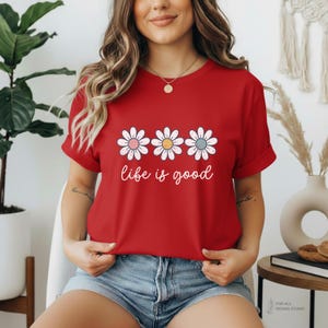 May include: A red t-shirt featuring three daisy flowers with pink, yellow, and blue centers. The words "life is good" are written in white script below the flowers. The shirt has short sleeves.