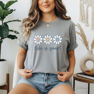 May include: A gray t-shirt featuring three daisy flowers with pink, yellow, and blue centers. The phrase "life is good" is written in a cursive font below the flowers. The shirt has rolled-up sleeves.