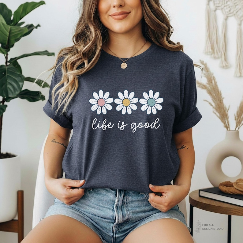 May include: A navy blue t-shirt featuring three daisy flowers with pink, yellow, and blue centers. The words "life is good" are printed below the flowers in a cursive font. The shirt is made of a soft material.
