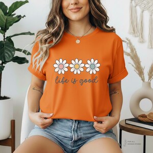 May include: Orange t-shirt with a daisy graphic and the phrase "life is good" in a script font. The daisy flowers have pink, yellow, and blue centers. The shirt has short sleeves and is made of a soft material.