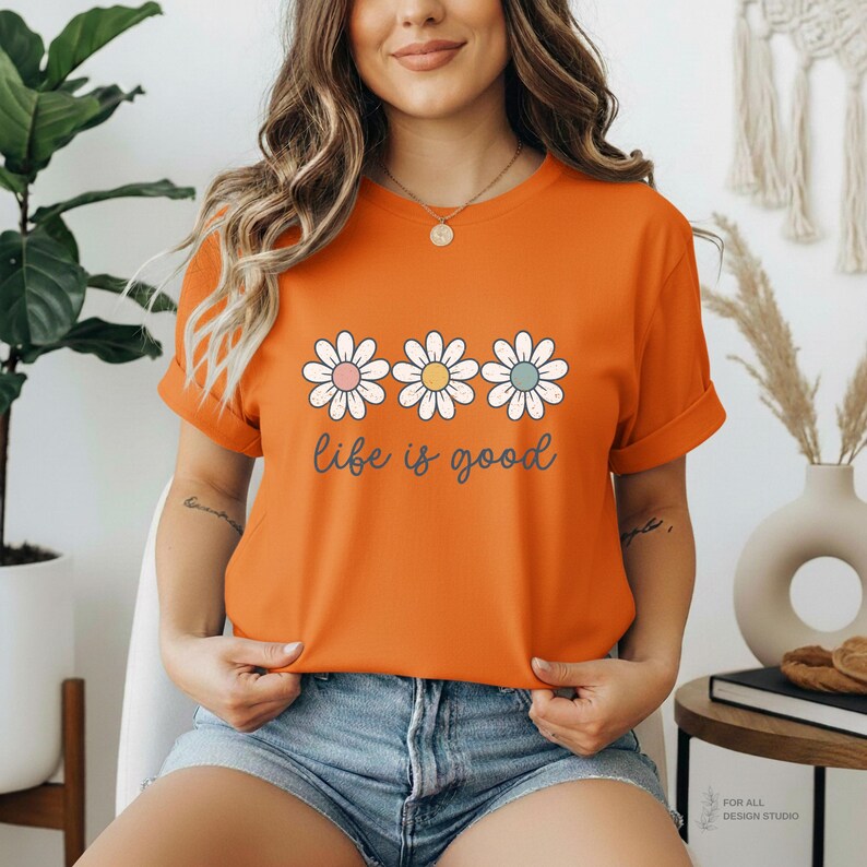 May include: Orange t-shirt with a daisy graphic and the phrase "life is good" in a script font. The daisy flowers have pink, yellow, and blue centers. The shirt has short sleeves and is made of a soft material.