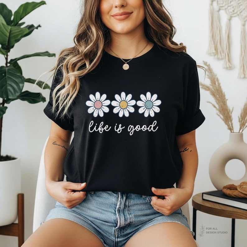 May include: Black t-shirt with a round neck featuring three daisy flowers with pastel-colored centers. The phrase "life is good" is written in a cursive font below the flowers. The shirt is worn with denim shorts.