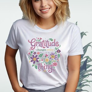 Könnte beinhalten: Weißes T-Shirt mit dem Schriftzug "Gratitude changes everything" in einem floralen Design. Das Design zeigt lila, rosa und gelbe Blumen und grüne Blätter. Das Shirt hat einen Rundhalsausschnitt.