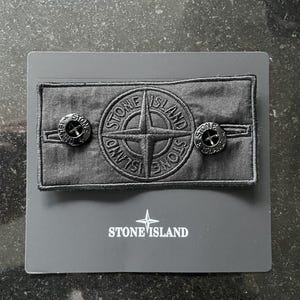Könnte beinhalten: Ein schwarzer Stone Island-Aufnäher mit einem Kompassrosen-Logo und zwei schwarzen Knöpfen. Der Aufnäher befindet sich auf einer grauen Karte mit dem Stone Island-Logo.