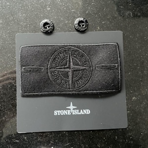 Op de afbeelding: Een zwarte Stone Island patch met het logo van het merk en twee zwarte knopen. De patch is rechthoekig met het Stone Island logo in het midden. De merknaam staat onderaan de kaart.