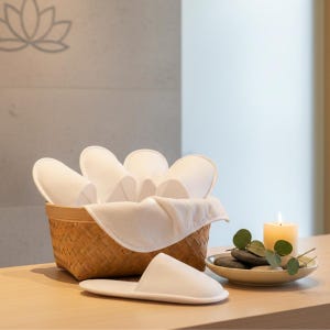 May include: A woven basket filled with white spa slippers, with one slipper in front. A lit candle, stones, and greenery sit on a small plate. The scene is set on a light wood surface, creating a relaxing spa-like atmosphere.