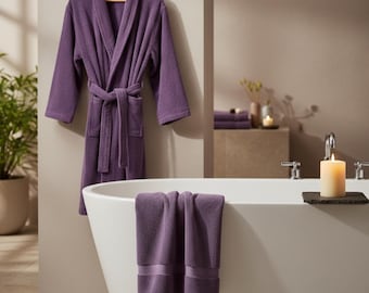 Puede incluir: Una bata de felpa morada cuelga de una percha de madera. Dos toallas moradas a juego están sobre el borde de una bañera blanca. Una vela encendida está sobre una bandeja de piedra negra.