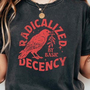 Op de afbeelding: Een antracietgrijs t-shirt met korte mouwen en een rode grafische print van een kraai die een takje vasthoudt. De tekst RADICALIZED BY BASIC DECENCY staat in een cirkel rond de vogel, geaccentueerd door kleine vierpuntige sterren.