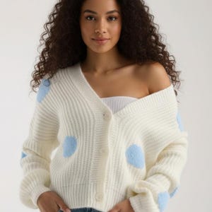 May include: Cream-colored cardigan sweater with light blue polka dots. The sweater has a V-neck, button closure, and long sleeves. The model is wearing blue jeans.