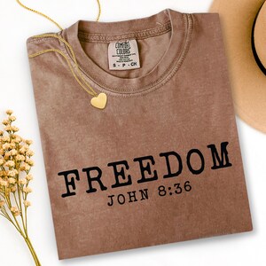 Könnte beinhalten: Ein braunes kurzärmeliges T-Shirt mit Rundhalsausschnitt und einem Comfort Colors Markenetikett. Das Shirt zeigt den schwarzen Schriftzug FREEDOM und JOHN 8:36 auf der Vorderseite. Eine goldfarbene Halskette mit herzförmigem Anhänger liegt auf dem Stoff.
