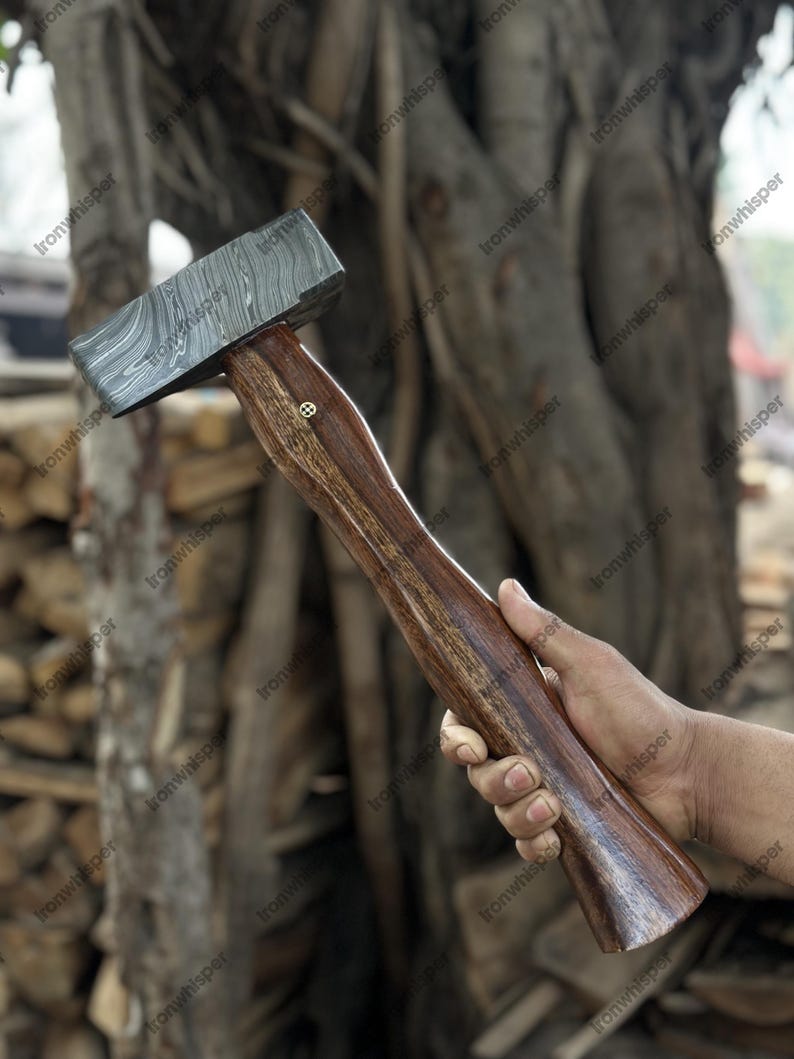 May include: A handcrafted hammer with a patterned steel head and a dark wooden handle. The hammer head has a unique, layered design. The handle is a rich brown color, and a small decorative element is visible near the head.