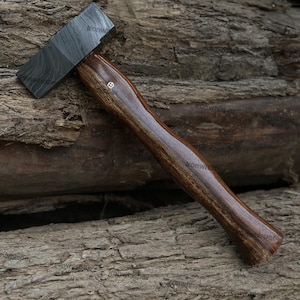 May include: A handcrafted hammer with a dark, patterned steel head and a polished, dark brown wooden handle. The handle has a smooth, ergonomic design and a small decorative element near the head. The hammer is displayed on a textured wooden surface.