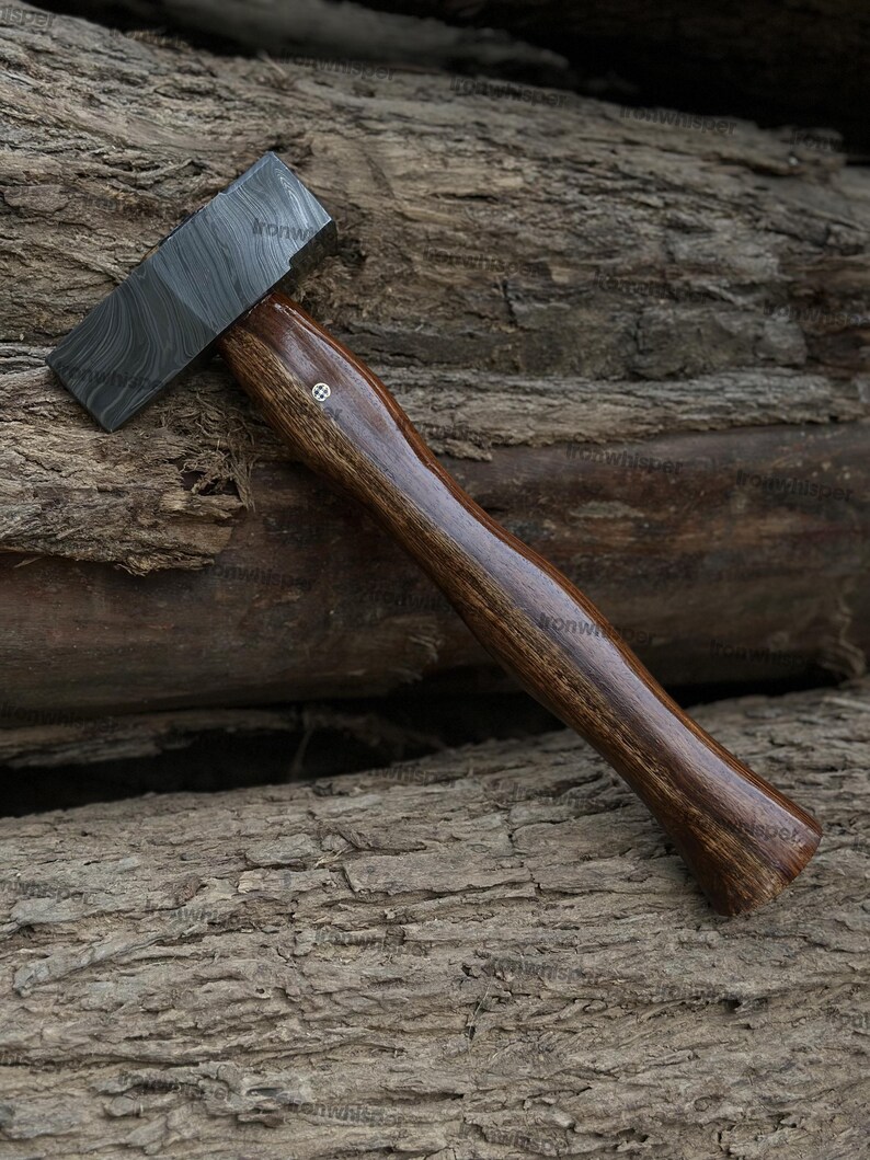 May include: A handcrafted hammer with a dark, patterned steel head and a polished, dark brown wooden handle. The handle has a smooth, ergonomic design and a small decorative element near the head. The hammer is displayed on a textured wooden surface.