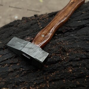 May include: A Damascus steel hammer with a dark wooden handle. The hammer head has a patterned design, and the handle is curved for grip. The hammer is resting on a dark, textured surface.