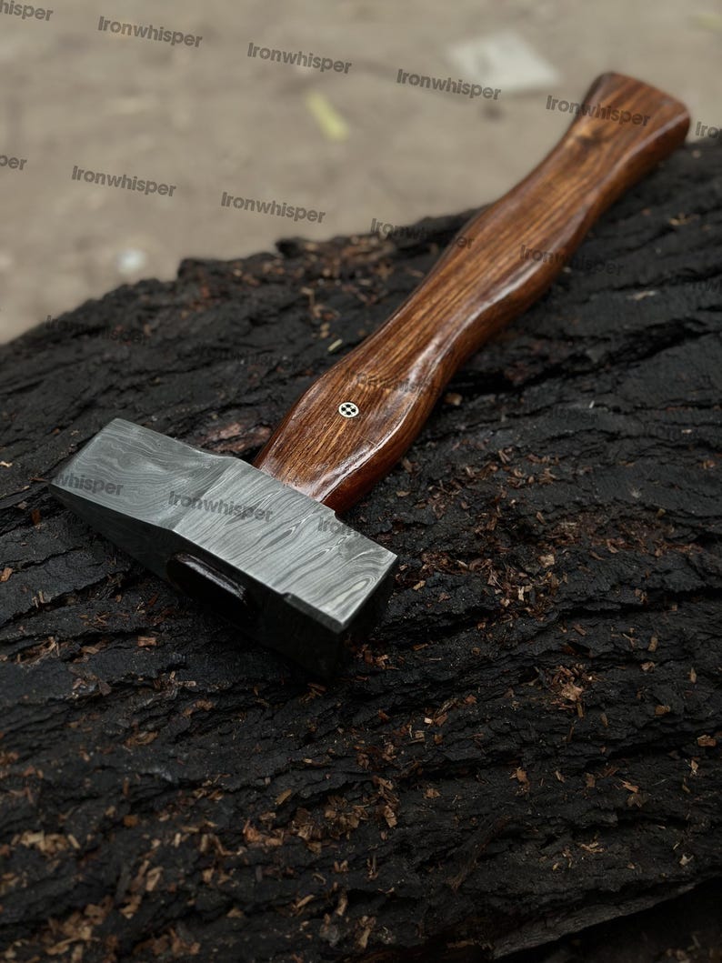 May include: A Damascus steel hammer with a dark wooden handle. The hammer head has a patterned design, and the handle is curved for grip. The hammer is resting on a dark, textured surface.