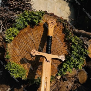 Puede incluir: Una espada de madera con empuñadura y hoja de color marrón claro, con empuñadura negra y grabados decorativos. La espada se exhibe sobre una superficie de madera con follaje verde, mostrando una estética rústica. El mango tiene el texto "Ironwhisper".