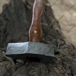 May include: A handcrafted hammer with a dark wooden handle and a patterned steel head. The hammer's head is a dark gray color with a unique swirling pattern. The handle is a rich brown color. A tool for woodworking or metalworking.
