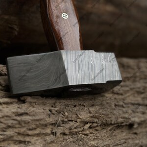 May include: Close-up of a blacksmith's hammer. The hammer features a dark, patterned metal head and a dark wooden handle. The handle has a decorative inlay near the head. The hammer rests on a textured, brown surface.