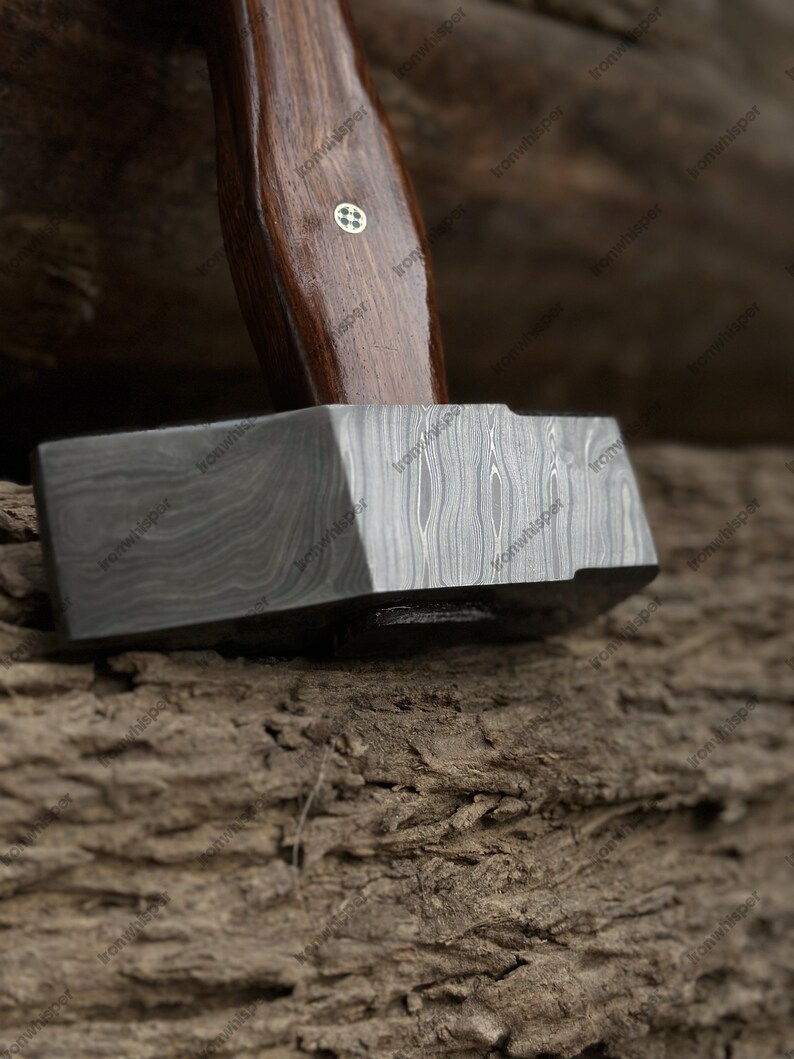 May include: Close-up of a blacksmith's hammer. The hammer features a dark, patterned metal head and a dark wooden handle. The handle has a decorative inlay near the head. The hammer rests on a textured, brown surface.