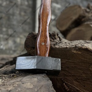 May include: A blacksmith's hammer with a dark, patterned steel head and a polished, dark wood handle. The handle has a slight hourglass shape and a small, decorative metal inlay. The hammer rests on a weathered wooden surface.