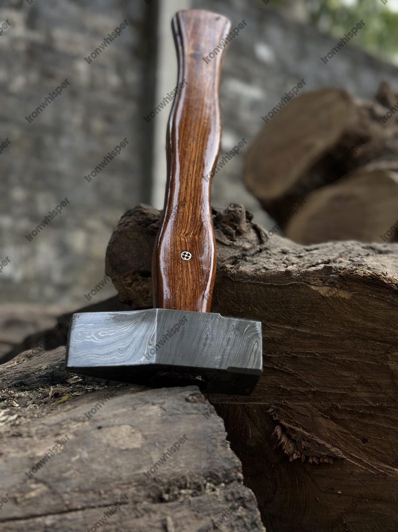 May include: A blacksmith's hammer with a dark, patterned steel head and a polished, dark wood handle. The handle has a slight hourglass shape and a small, decorative metal inlay. The hammer rests on a weathered wooden surface.