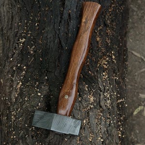 May include: A handcrafted axe with a dark wood handle and a patterned steel head. The handle is curved and polished, with a brass accent. The axe is resting on a textured, dark brown tree trunk.