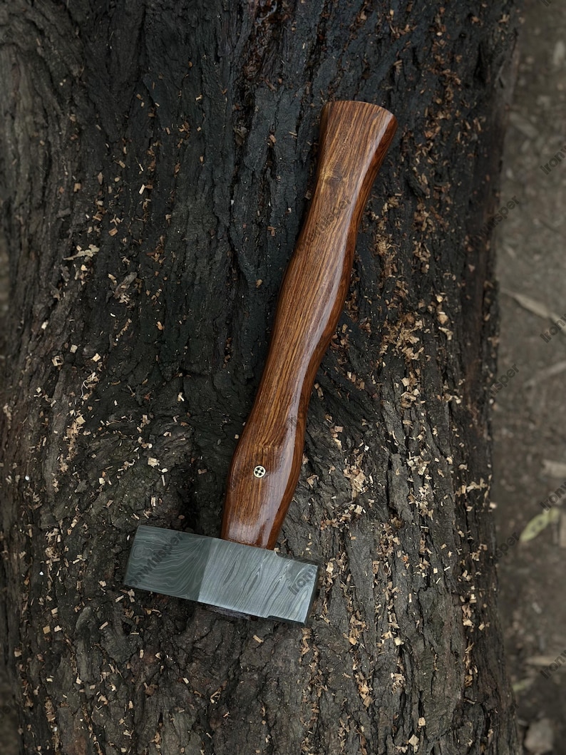 May include: A handcrafted axe with a dark wood handle and a patterned steel head. The handle is curved and polished, with a brass accent. The axe is resting on a textured, dark brown tree trunk.
