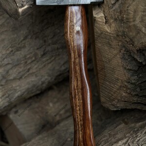 May include: A handcrafted hammer with a patterned steel head and a dark, polished wooden handle. The handle features a decorative emblem. The hammer is set against a backdrop of stacked wood.
