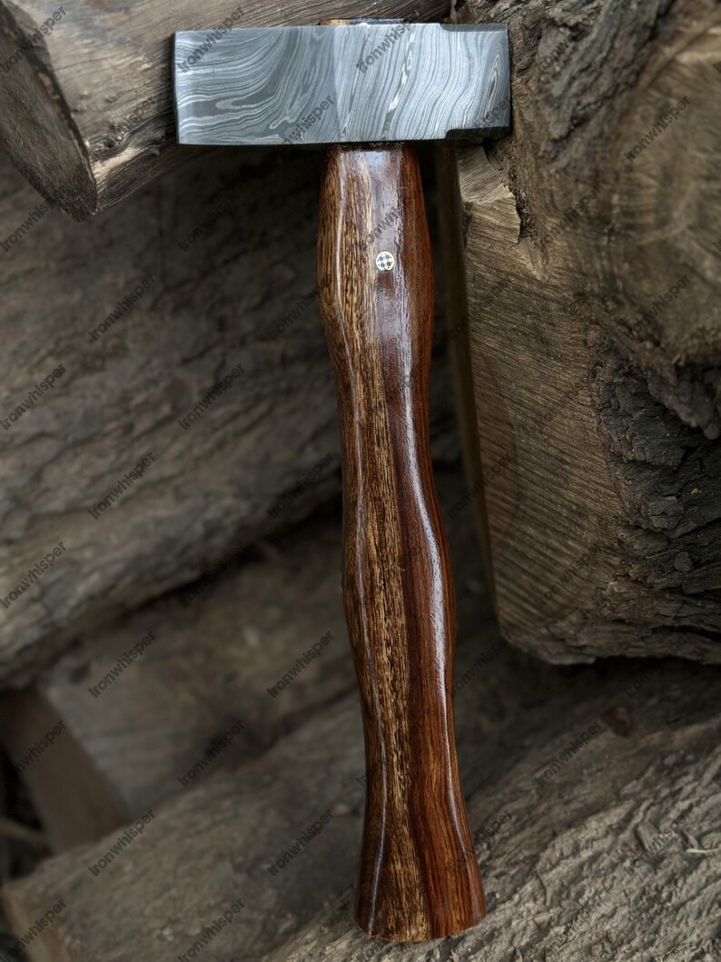 May include: A handcrafted hammer with a patterned steel head and a dark, polished wooden handle. The handle features a decorative emblem. The hammer is set against a backdrop of stacked wood.
