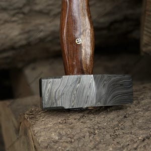 May include: A close-up of a hammer with a dark, patterned metal head and a wooden handle. The handle is a rich brown color with a small, gold-colored emblem. The hammer rests on a rough, textured surface.