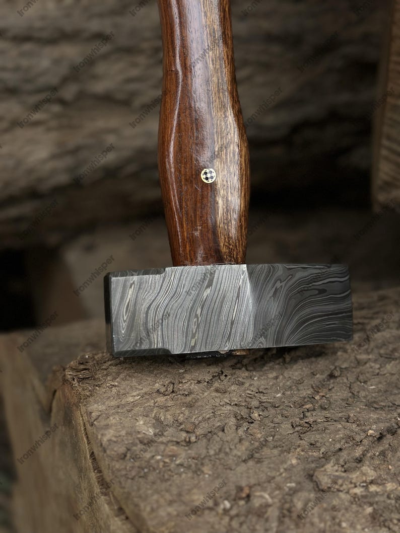 May include: A close-up of a hammer with a dark, patterned metal head and a wooden handle. The handle is a rich brown color with a small, gold-colored emblem. The hammer rests on a rough, textured surface.