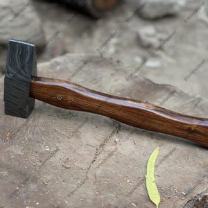 May include: A blacksmith's hammer with a dark, patterned steel head and a polished wooden handle. The hammer is resting on a weathered wooden surface, showcasing the tool's craftsmanship and design. A leaf lies nearby.