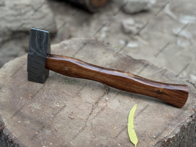 May include: A blacksmith's hammer with a dark, patterned steel head and a polished wooden handle. The hammer is resting on a weathered wooden surface, showcasing the tool's craftsmanship and design. A leaf lies nearby.