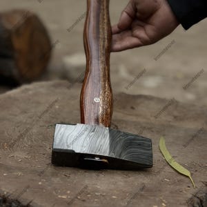 May include: A handcrafted hammer with a patterned steel head and a wooden handle. The handle is a dark brown color with lighter brown wood grain. The hammer is resting on a wooden surface.