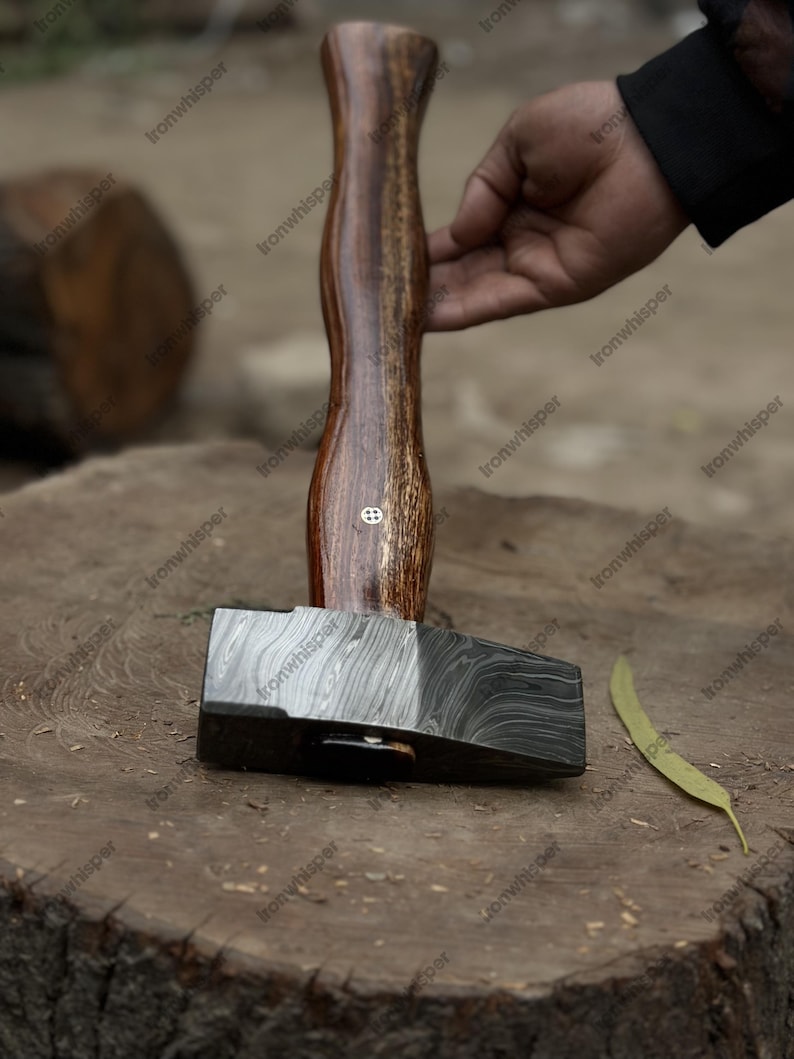 May include: A handcrafted hammer with a patterned steel head and a wooden handle. The handle is a dark brown color with lighter brown wood grain. The hammer is resting on a wooden surface.