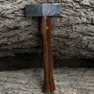May include: A hammer with a patterned steel head and a dark wooden handle. The handle has a curved design and a small, decorative brass element. The hammer is displayed against a textured, brown background.