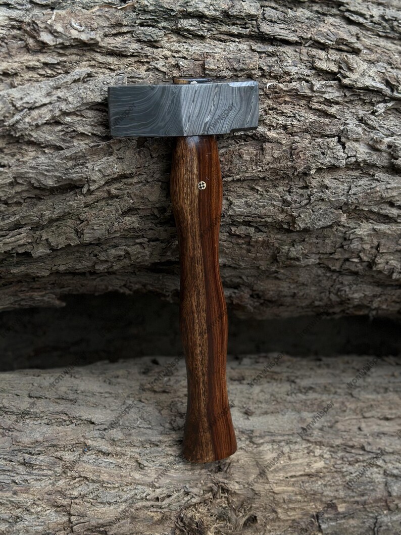 May include: A hammer with a patterned steel head and a dark wooden handle. The handle has a curved design and a small, decorative brass element. The hammer is displayed against a textured, brown background.