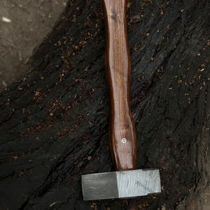May include: A handcrafted hammer with a dark wooden handle and a patterned steel head. The handle is a rich brown color, and the head has a contrasting silver and dark gray pattern. The hammer is designed for woodworking or metalworking.