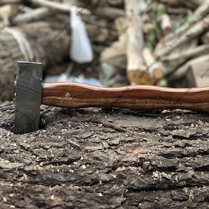 May include: A handcrafted hammer with a dark, textured steel head and a curved, polished wooden handle. The hammer is resting on a textured, brown tree trunk, showcasing its craftsmanship and design. The handle has a small brass pin.