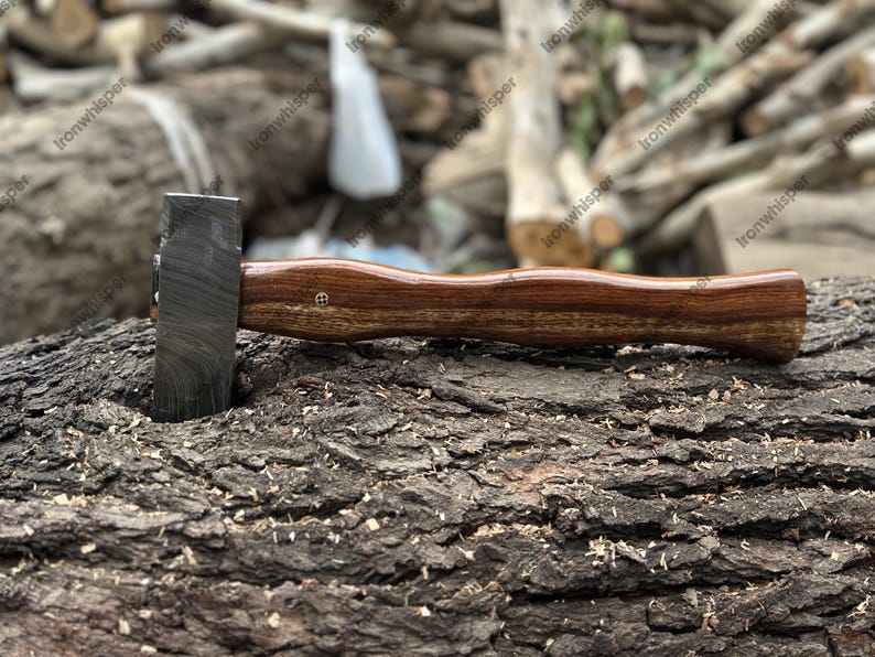 May include: A handcrafted hammer with a dark, textured steel head and a curved, polished wooden handle. The hammer is resting on a textured, brown tree trunk, showcasing its craftsmanship and design. The handle has a small brass pin.