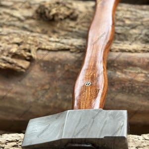May include: A handcrafted axe with a dark wooden handle and a patterned steel head. The handle has a curved design and is attached to the axe head with a small metal pin. The axe head has a textured, layered appearance.