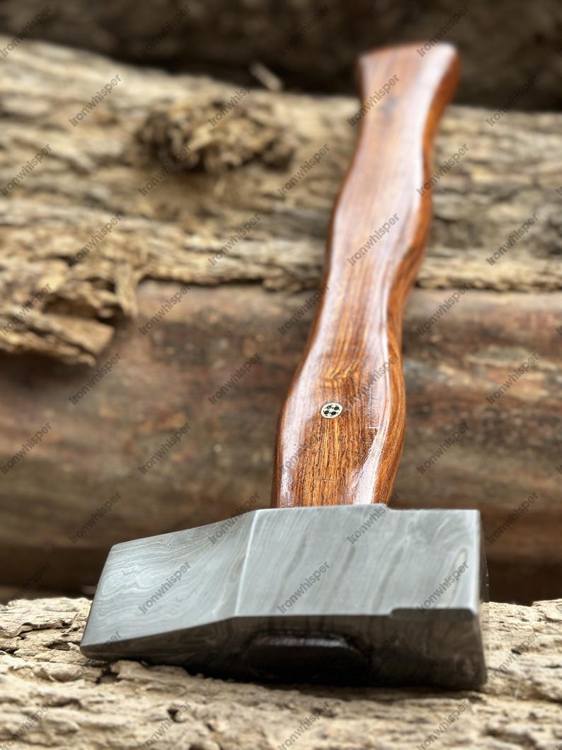 May include: A handcrafted axe with a dark wooden handle and a patterned steel head. The handle has a curved design and is attached to the axe head with a small metal pin. The axe head has a textured, layered appearance.