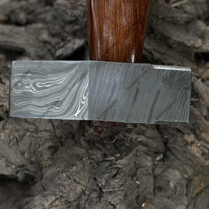 May include: A close-up of a hammer with a patterned steel head and a wooden handle. The handle is a dark brown color with a small decorative element. The hammer head has a unique, swirling pattern.