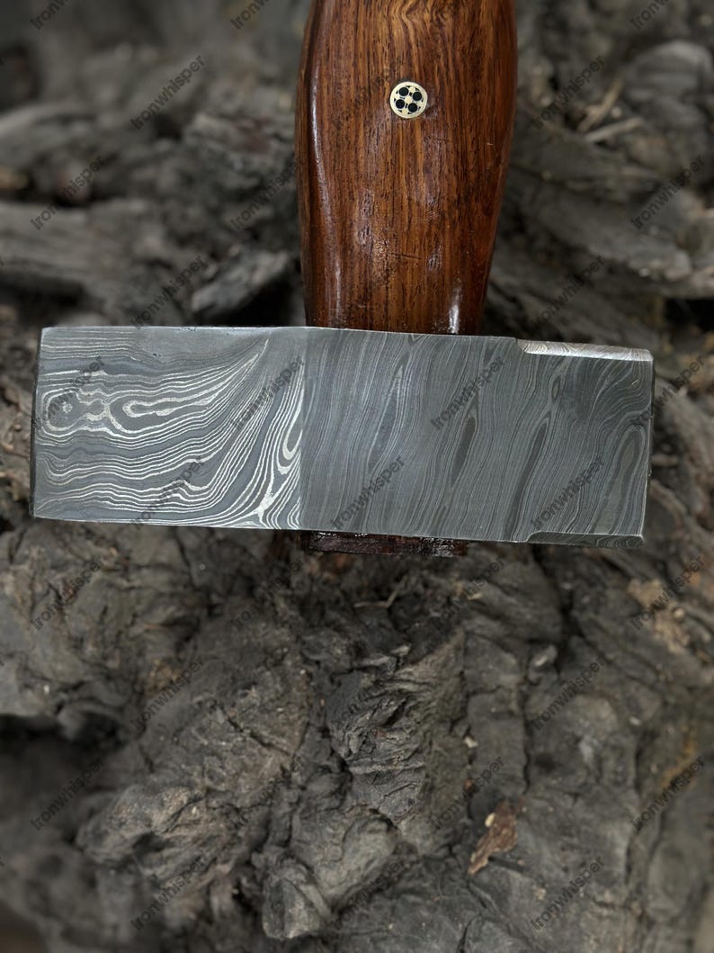 May include: A close-up of a hammer with a patterned steel head and a wooden handle. The handle is a dark brown color with a small decorative element. The hammer head has a unique, swirling pattern.